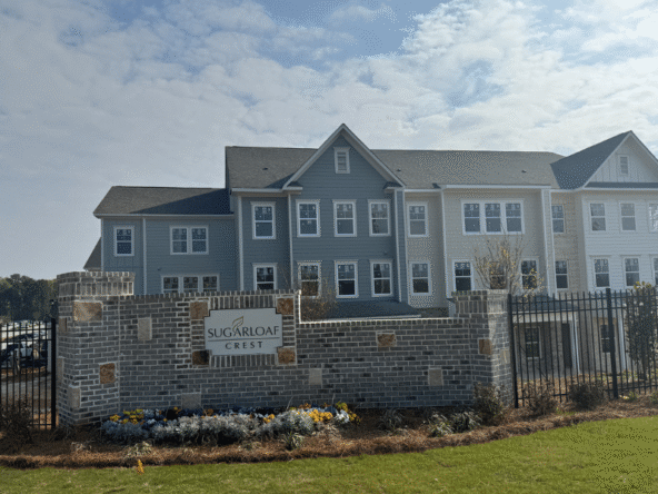 Two-story apartment complex with blue and gray siding, seen behind a brick entrance wall and black metal fence, sign reads Sugarloaf Crest, landscaped with flowers.