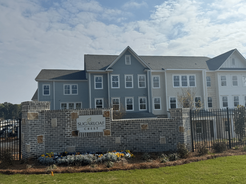 Two-story apartment complex with blue and gray siding, seen behind a brick entrance wall and black metal fence, sign reads Sugarloaf Crest, landscaped with flowers.